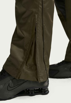 Olivengrønne bukser med sidelukning med lynlås, delvist åben, over sorte Nike-sneakers med forårslignende såldesign.