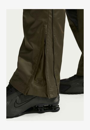 Pantaloni verde oliva con dettaglio di cerniera laterale parzialmente aperta sopra sneakers nere Nike con suola dal design a molla.