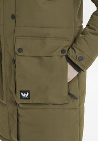 Olivgrön isolerad parka med svarta tryckknappar, två framfickor, ribbade ärmslut och en logotyp-patch med texten "Whistler."