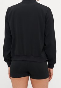 Black bomber jacket with a ribbed collar and cuffs, featuring a smooth texture. Cut with a cropped design, sitting above a pair of black shorts.