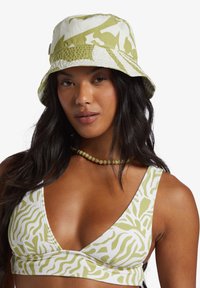 Frau mit langen dunklen Haaren, die einen weißen und hellgrünen gemusterten Bucket Hat, ein passendes Bralette und eine Perlenkette vor einem weißen Hintergrund trägt.