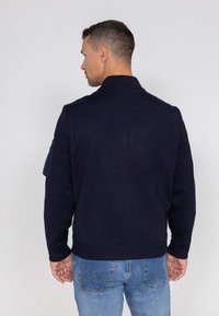Navyblauwe gebreide cardigan met kraag, geribde zoom en textielstructuur. Bevat een patch op de bovenste linkermouw.