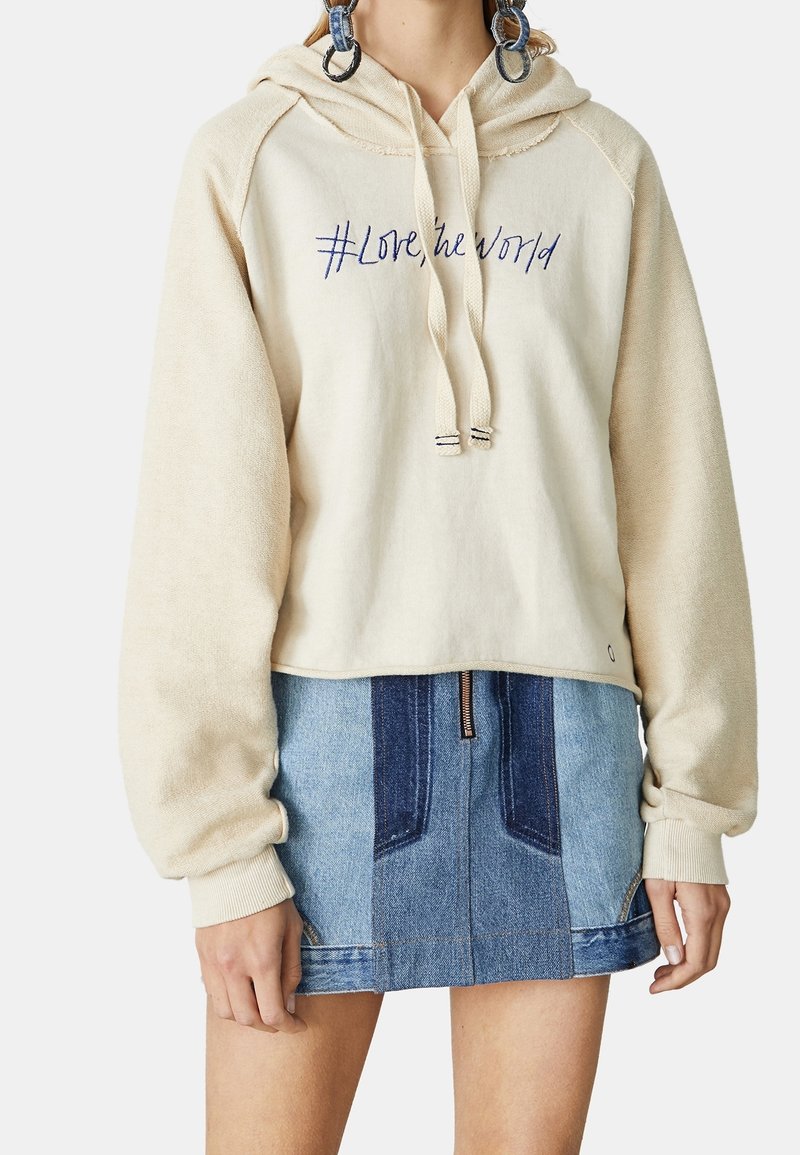Sweat à capuche beige avec le texte "#Love the World" associé à une mini-jupe en jean patchwork bleue et de grandes boucles d'oreilles créoles sur une personne debout.