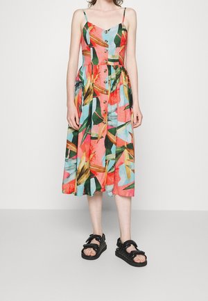 Femme portant une robe midi sans manches à boutons sur le devant avec des motifs colorés de feuilles tropicales et des sandales noires à bout ouvert avec des lanières.