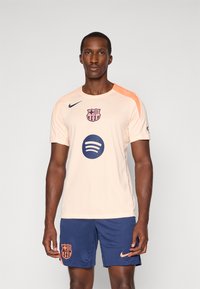 Licht perzikkleurig voetbalshirt met marineblauwe accenten, voorzien van het Barcelona-embleem en het Spotify-logo. Gecombineerd met marineblauwe shorts, Nike-branding zichtbaar.