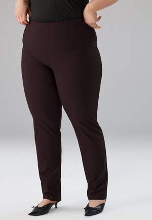 Kurvige Person trägt eng anliegende dunkelbraune Hose und schwarze spitze Kitten-Heel-Schuhe, steht vor einem schlichten grauen Hintergrund.