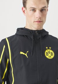 Chaqueta negra con cremallera y capucha, con acentos amarillos, un logo de Puma y un logo circular amarillo de club que tiene "BVB 09" y estrellas.