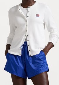 Osoba v bílém sáčku s knoflíky, na kterém je americká vlajka a logo "Polo", pod kterým má bílý top, doplněná jasně modrými šortkami na stažení.