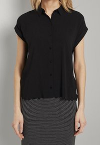 Donna che indossa una camicia nera a maniche corte con bottoni abbinata a una gonna a righe orizzontali bianco e nera, in piedi davanti a uno sfondo semplice.