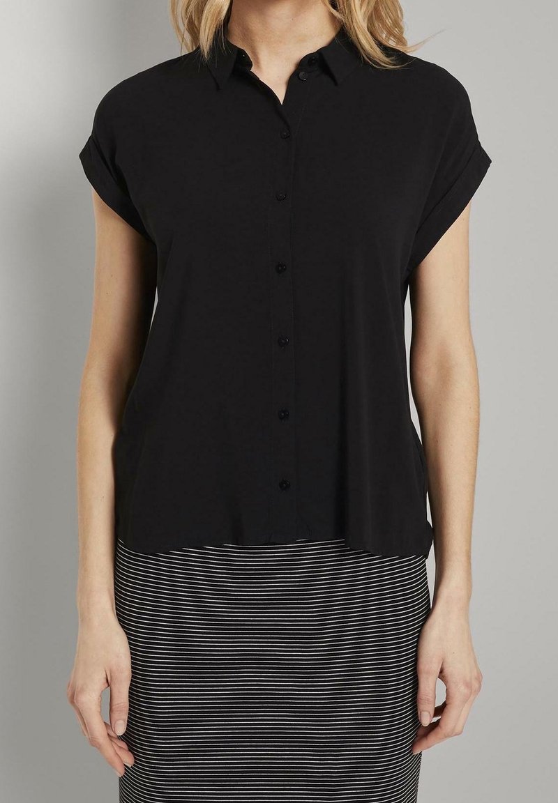 Donna che indossa una camicia nera a maniche corte con bottoni abbinata a una gonna a righe orizzontali bianco e nera, in piedi davanti a uno sfondo semplice.