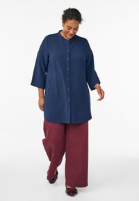 Camicia blu navy con tessuto testurizzato e colletto, abbinata a pantaloni larghi bordeaux e scarpe scure.