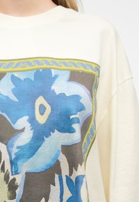 Μεγάλο μπλε λουλουδένιο μοτίβο με κίτρινες και γκρι πινελιές, περιβεβλημένο από κίτρινο περίγραμμα, στο μπροστινό μέρος ενός μπλουζιού με μακριά μανίκια σε κρεμ απόχρωση.