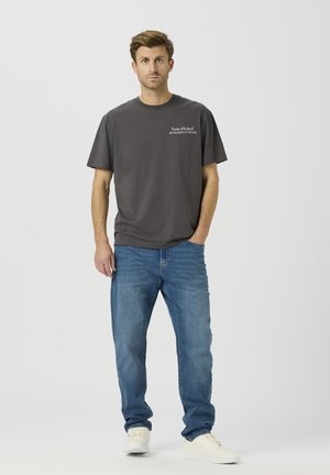 Mørkegrå bomuld t-shirt med korte ærmer, med hvid tekst på venstre bryst. Kombineret med blå cowboybukser og hvide sneakers.