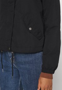 Svart jacka med knappsläppficka och dragsnören, bärs över blå jeans, visar en brun ribbad manschett och en hand vid sidan.