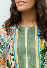Blouse multicolore à motif floral sur fond vert, avec des rayures verticales bleues, jaunes et blanches ; tissu léger.