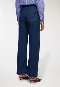 Jeans in denim blu scuro a vita alta e gamba larga, con cuciture arancioni a contrasto e due tasche posteriori, abbinati a mule borgogna.