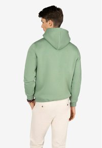 Sudadera con capucha verde de corte relajado, que presenta un bolsillo canguro y puños acanalados, combinada con pantalones ligeros de color beige.