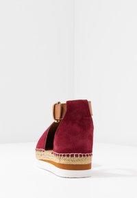 Sandale compensée en suède bordeaux avec une bride de cheville beige, semelle en jute tressé et semelle extérieure en caoutchouc blanc. L'arrière présente un détail logo en relief.