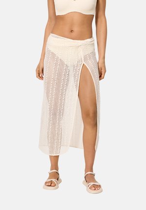 Mujer con una falda cruzada de crochet crema con una abertura alta, combinada con un bikini a juego y sandalias, sobre un fondo liso.