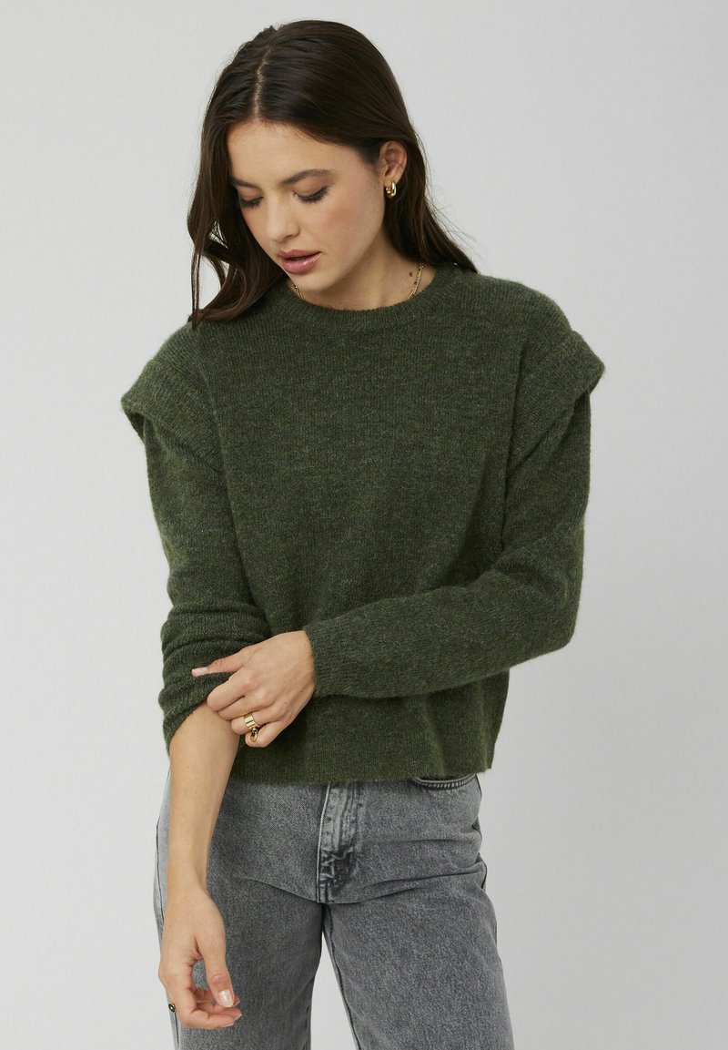 Pull en maille verte avec des épaules tombantes, des poignets côtelés et une coupe décontractée, associé à un jean gris taille haute.