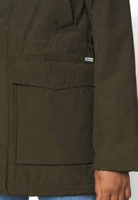 Giacca verde scuro con una texture liscia, grandi tasche anteriori e una piccola etichetta con logo sulla tasca sinistra.