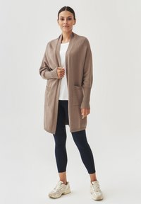 Long cardigan ouvert en marron clair, fabriqué en matériau tricoté doux. Il présente des poignets côtelés et deux poches avant. Porté sur un haut blanc.