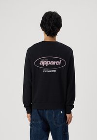 Zwarte sweatshirt met lange mouwen, geribbelde manchetten en zoom, met een groot roze logo op de achterkant. Draag het over blauwe denim broek.