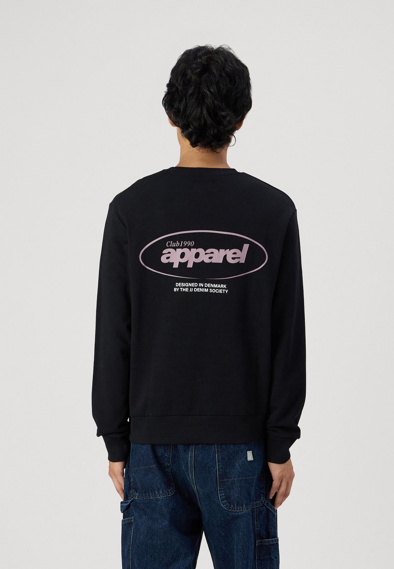 Sudadera negra de manga larga, con puños y dobladillo acanalados, que presenta un gran logo rosa en la parte posterior. Llevada sobre pantalones de mezclilla azul.