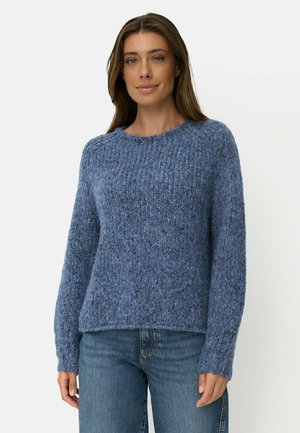 Kvinde iført en tekstureret blå strikket sweater og blå jeans, stående mod en ensfarvet lys baggrund, vendt fremad med et let smil.