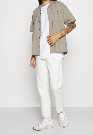 Persona con camisa beige de manga corta abotonada sobre una camiseta blanca, pantalones blancos y zapatillas blancas con detalles en rosa, de pie en el interior.