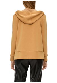 Sweat à capuche marron avec des poignets et un ourlet côtelés. Texture douce, coupe décontractée. Porté avec un pantalon en cuir noir, affichant un minimum de détails de couture.