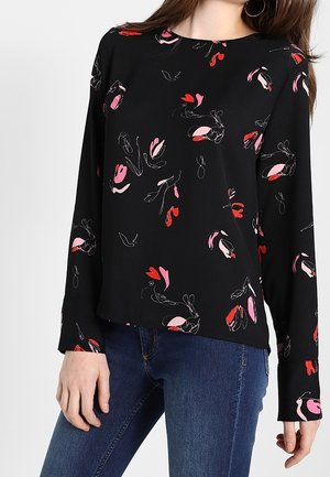 Femme portant un chemisier noir à manches longues avec des motifs floraux abstraits rouges et roses et un jean bleu, debout avec les mains détendues.