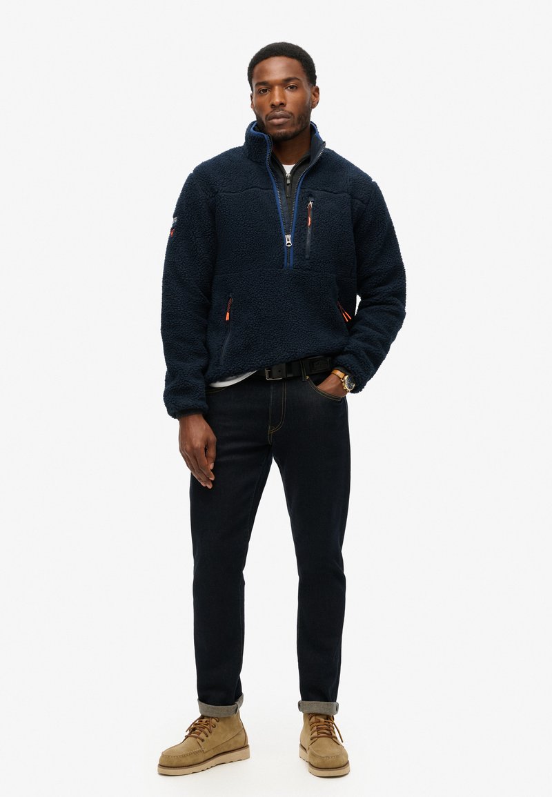 Marineblauwe fleece pullover met een halve rits, twee zijzakken, contrasterende oranje hardware en een relaxte pasvorm, gecombineerd met donkere jeans en beige laarzen.