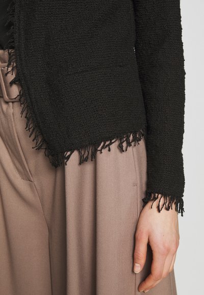 Veste noire texturée avec des franges le long de l'ourlet et des manches, portée sur un pantalon beige taille haute. Vue en gros plan.