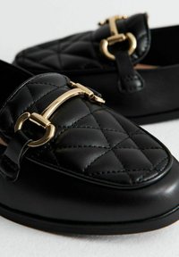 Zwarte gequiltte leren instappers met een gold-tone hardware accent. Gladde textuur, ronde neus en subtiele stikdetails.