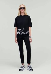 T-shirt preto de manga curta com o logótipo branco "Karl", combinado com jeans skinny pretos e sapatilhas chunky preto e branco. Modelo com óculos de sol.
