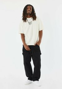 Camiseta de manga corta color crema con estampado gráfico, combinada con pantalones cargo negros de corte holgado y zapatillas blancas. Diseño simple, tejido ligero.