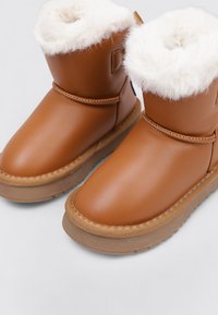 Botines sintéticos marrones con un forro blanco y esponjoso de piel sintética, con una suela de goma texturizada de color marrón claro y detalles simples de costura.