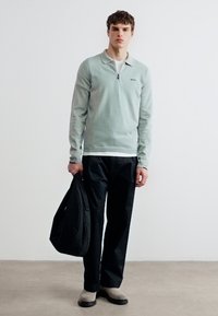 Νεαρός άνδρας όρθιος, φοράει ελαφρύ πράσινο πουλόβερ με φερμουάρ, μαύρο παντελόνι, μπεζ παπούτσια, κρατώντας μαύρη τσάντα στο αριστερό χέρι μπροστά σε απλό φόντο.