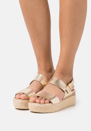 Sandales métalliques dorées avec deux larges brides, une semelle plateforme en jute tressé, et une bride à la cheville avec boucle, présentant une texture lisse.