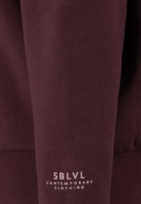Bordeaux sweatshirt met een geribbelde zoom en manchetten, met een witte bedrukte logo dat "SBLVL CONTEMPORARY CLOTHING" op de mouw leest.