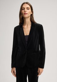Donna che indossa un blazer di velluto nero sopra una maglietta nera, in piedi davanti a uno sfondo grigio chiaro uniforme, rivolta verso l'avanti.