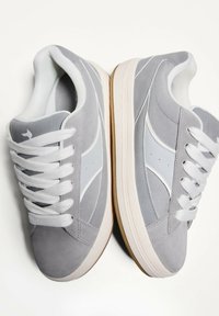 Sneakers grigi con una parte superiore in suede testurizzato e accenti sintetici lisci. Presentano lacci bianchi e una suola in gomma di colore chiaro.
