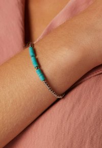 Kralenarmband met turquoise cilinderkralen en zilveren ronde kralen op een getextureerde arm, tegen een roze stoffen achtergrond.