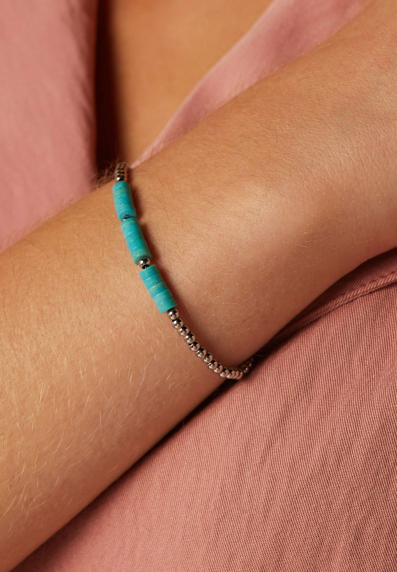 Kralenarmband met turquoise cilinderkralen en zilveren ronde kralen op een getextureerde arm, tegen een roze stoffen achtergrond.