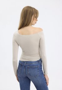 Langärmliges, schulterfreies beige Oberteil, kombiniert mit hochtaillierten blauen Jeans, die eine glatte Textur und einen taillierten Schnitt zeigen.