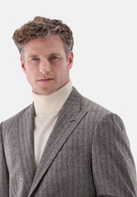 Brauner Fischgrät-Blazer mit Reverskragen, über einem crèmefarbenen Rollkragenpullover getragen. Verfügt über strukturierte Stoffqualität und maßgeschneiderten Schnitt.