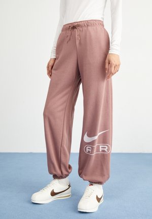 Personne portant un pantalon de survêtement mauve avec le logo blanc Nike Air et des baskets blanches Nike, debout sur un sol bleu clair.