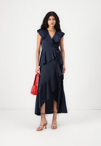 Vestido azul-marinho com mangas franzidas e bainha em cascata, combinado com sandálias beijas de tiras e uma pequena bolsa vermelha. Textura suave.
