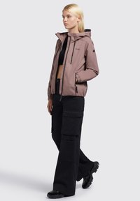 Helle rosa leichte Jacke mit Kapuze, mit Reißverschlusstaschen, elastischen Bündchen, kombiniert mit schwarzen Cargo-Hosen und schwarzem Schuhwerk.